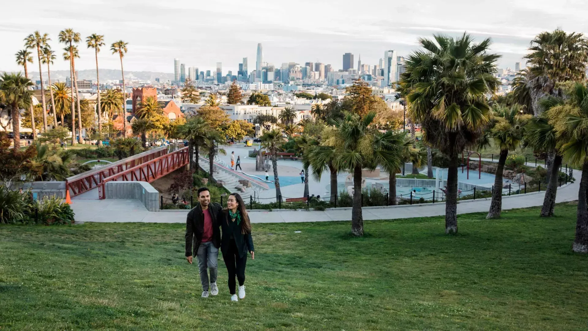 한 커플이 Dolores Park 과 샌프란시스코 스카이라인을 뒤로 한 채 카메라를 향해 걸어가고 있습니다.