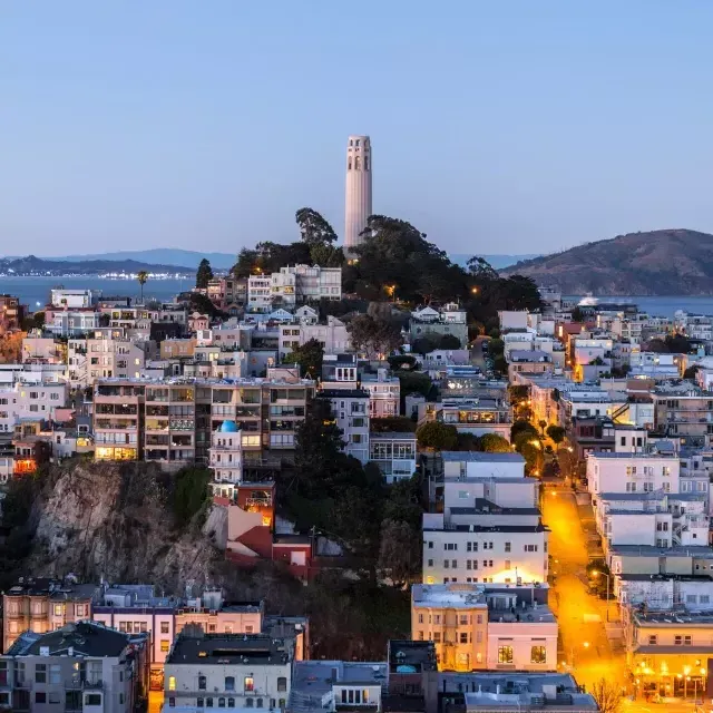 Coit Tower em São Francisco, ao anoitecer, com ruas iluminadas à sua frente e a Baía de São Francisco atrás.